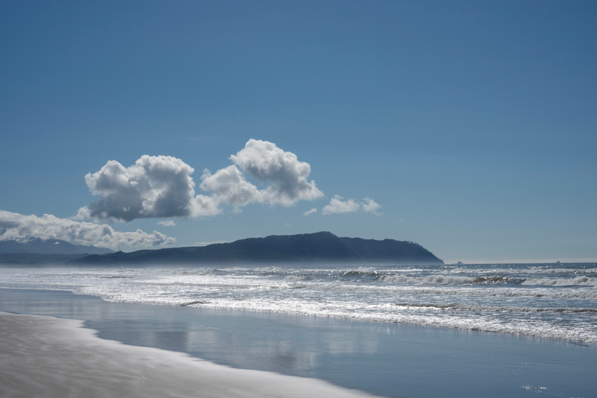 Waves rolling onto a broad beach beneath clouds and a wooded coastal headland.
