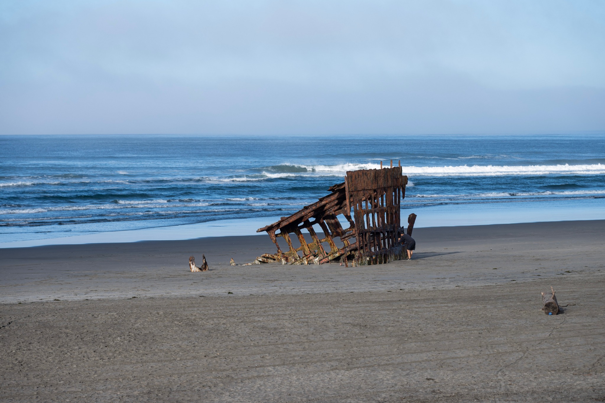 Rusted shipwreck remains on a broad sandy beach with rolling ocean waves beyond.
